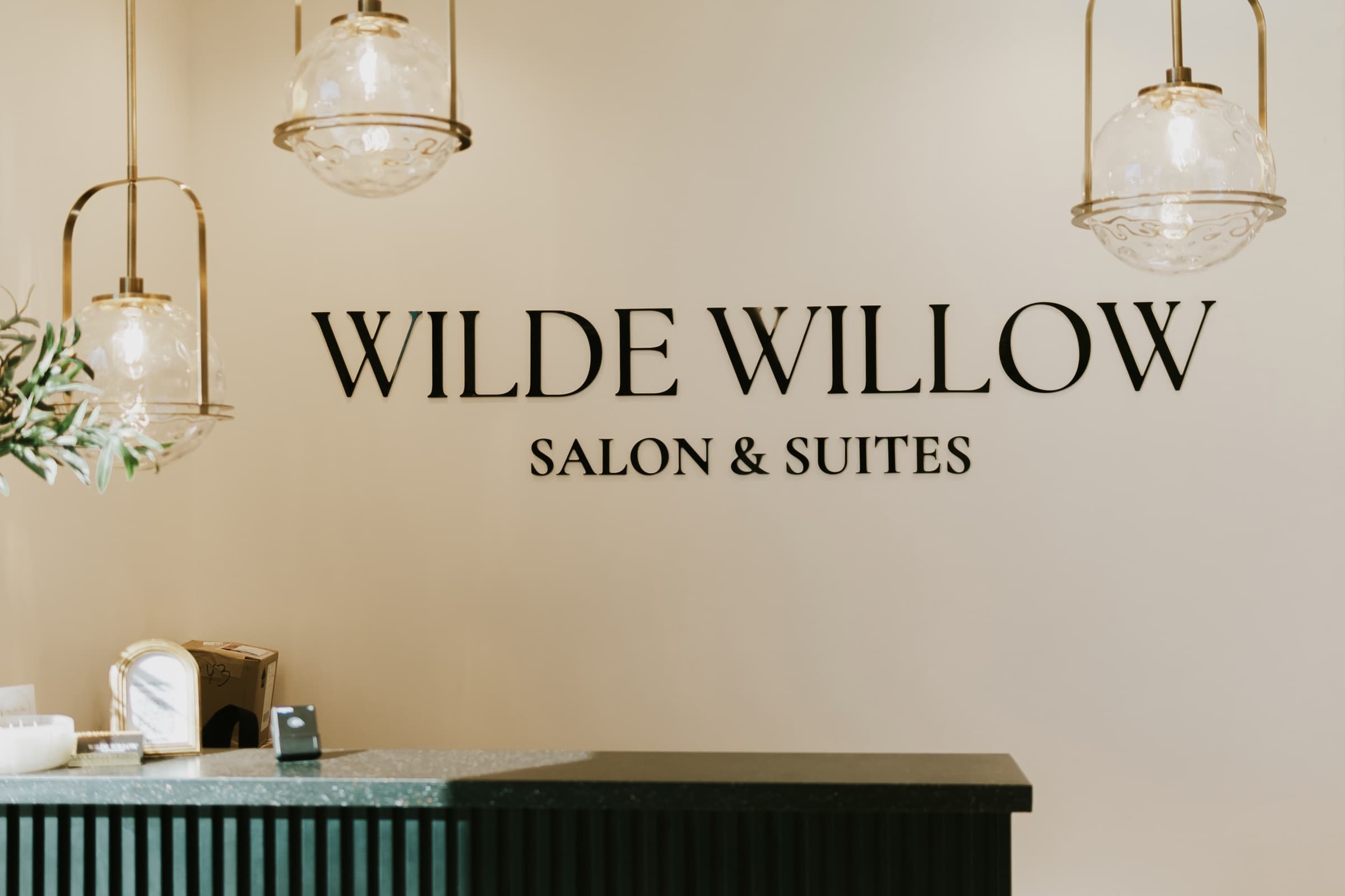 The Wilde Willow Salon & Suites reception wall, lit by three brass globe pendants.