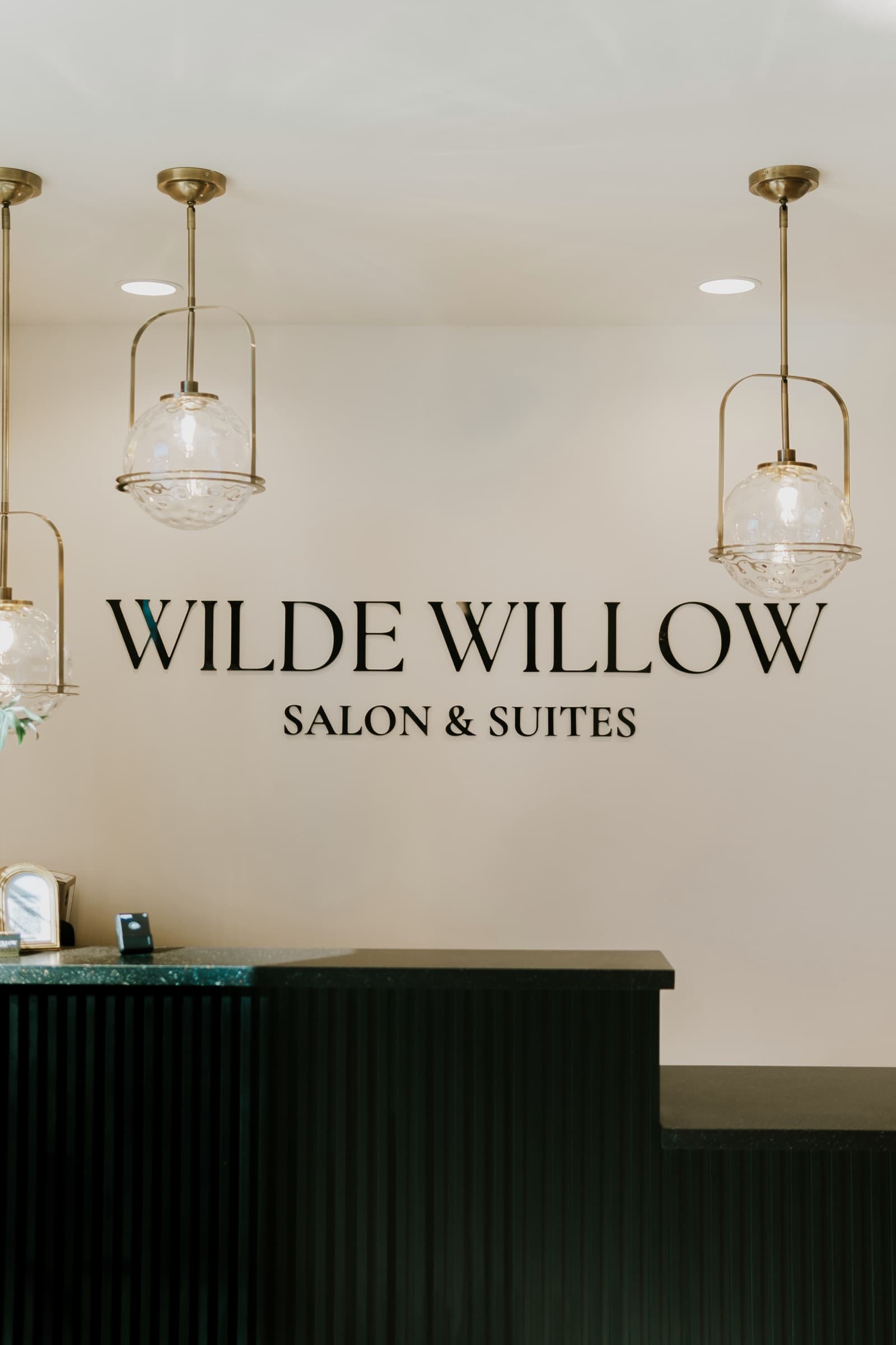 The reception desk beneath the Wilde Willow Salon & Suites wordmark, lit by brass globe pendants.